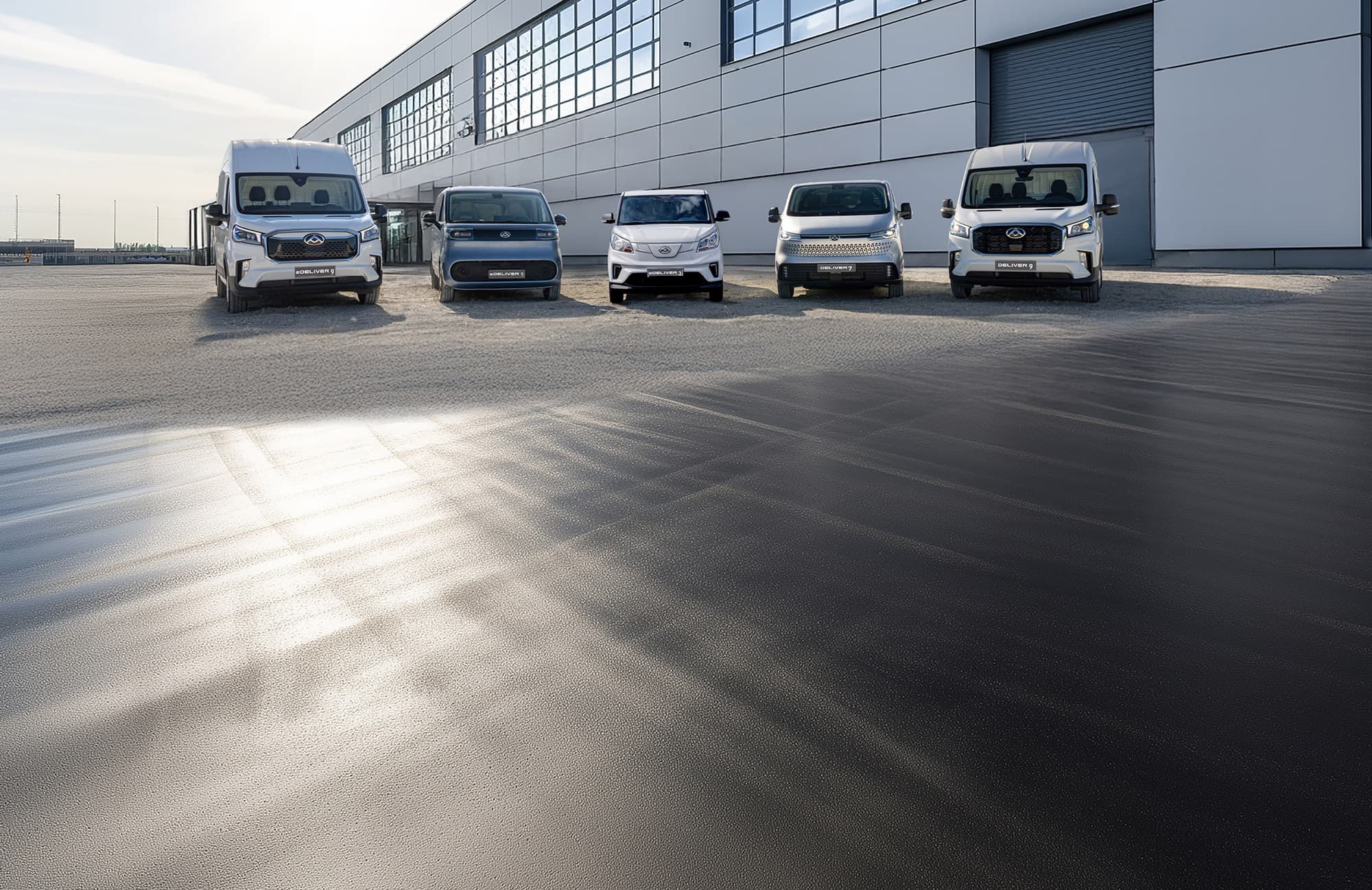 Five Maxus vans are parked in a row on a paved surface near a modern industrial building under a clear sky.