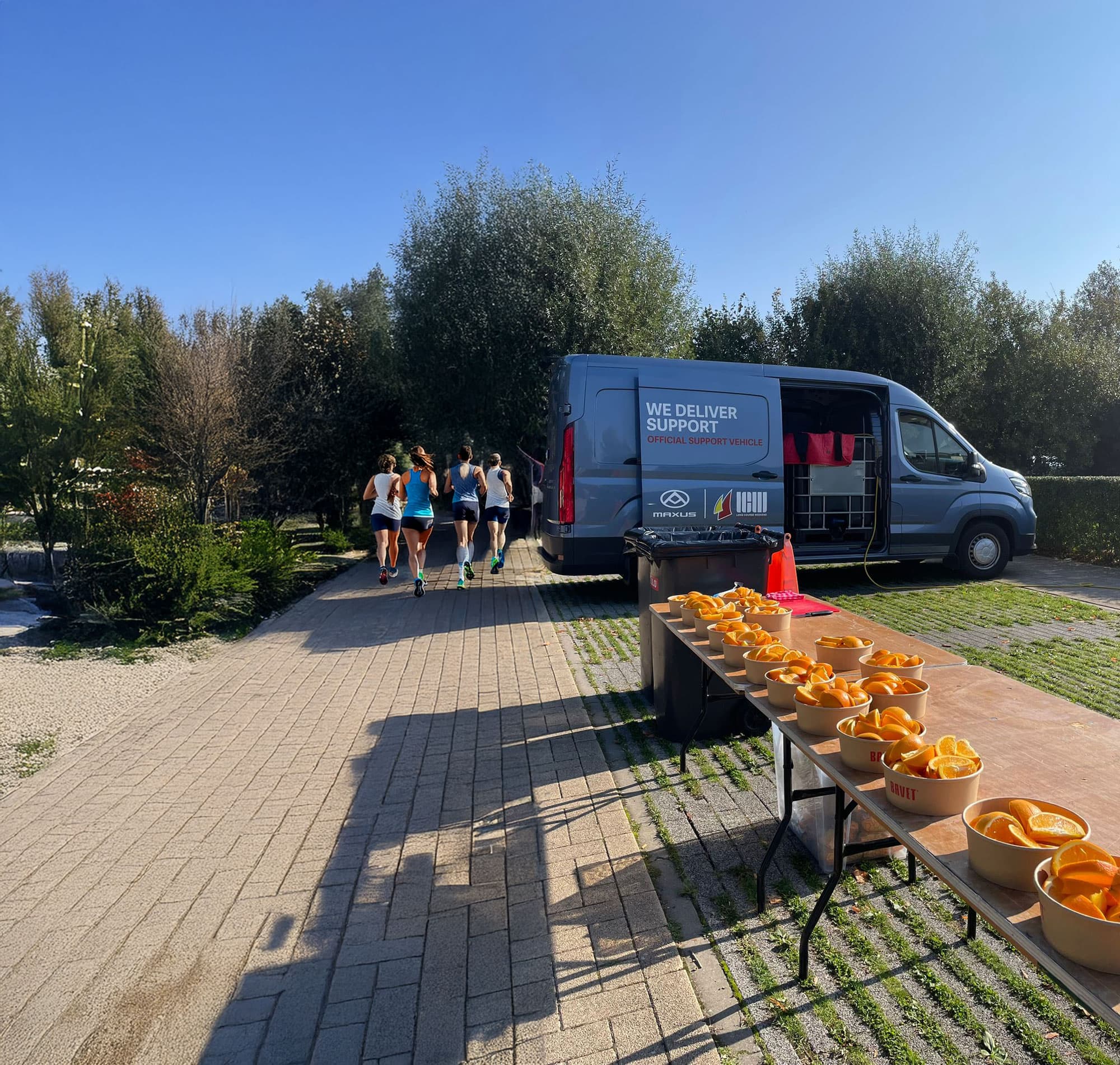 A group of runners jogs past a van and tables with bowls of sliced oranges on a sunny day, surrounded by trees.
