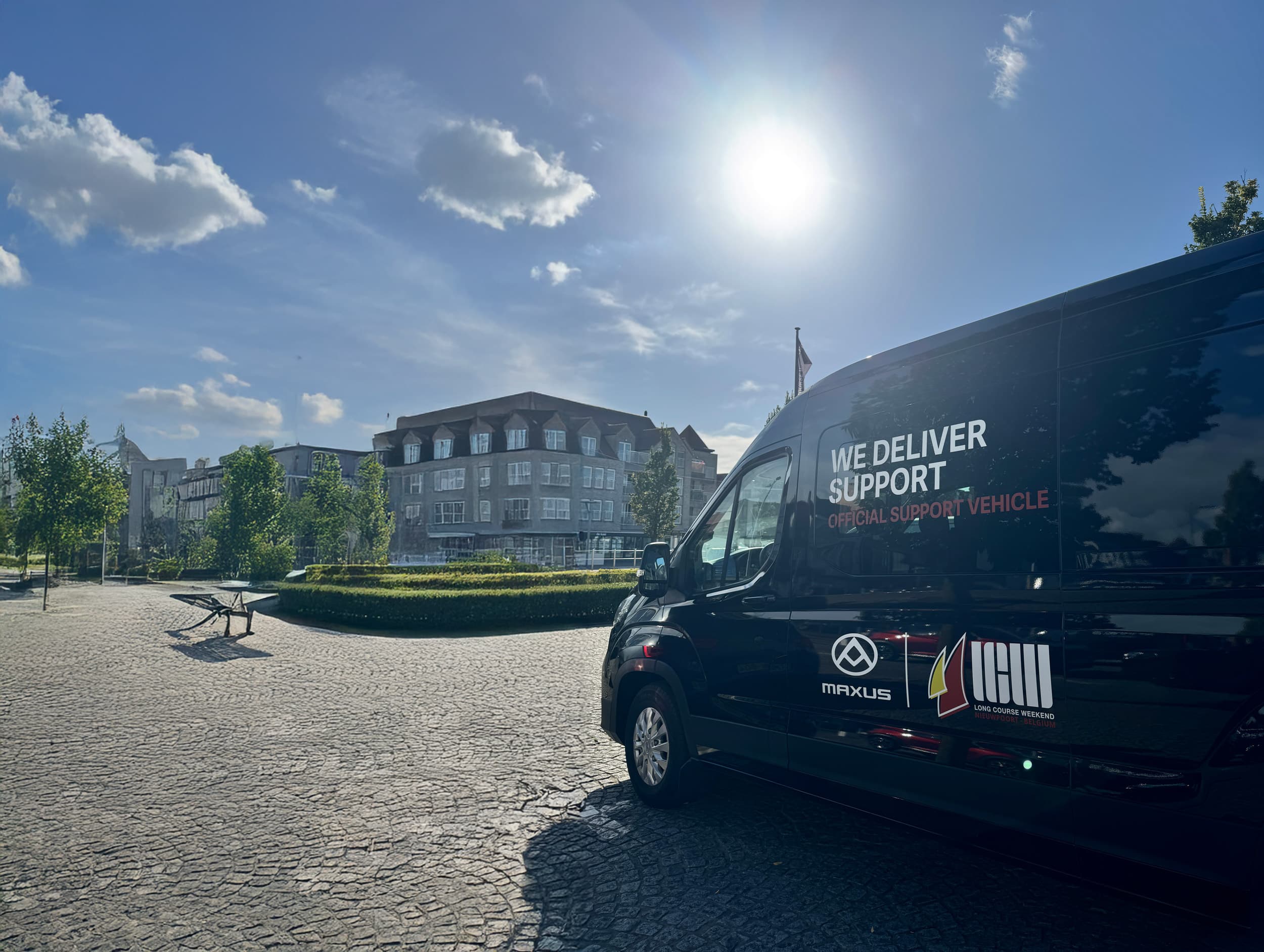 A black support van is parked on a sunlit cobblestone plaza with a historic building and trees in the background.