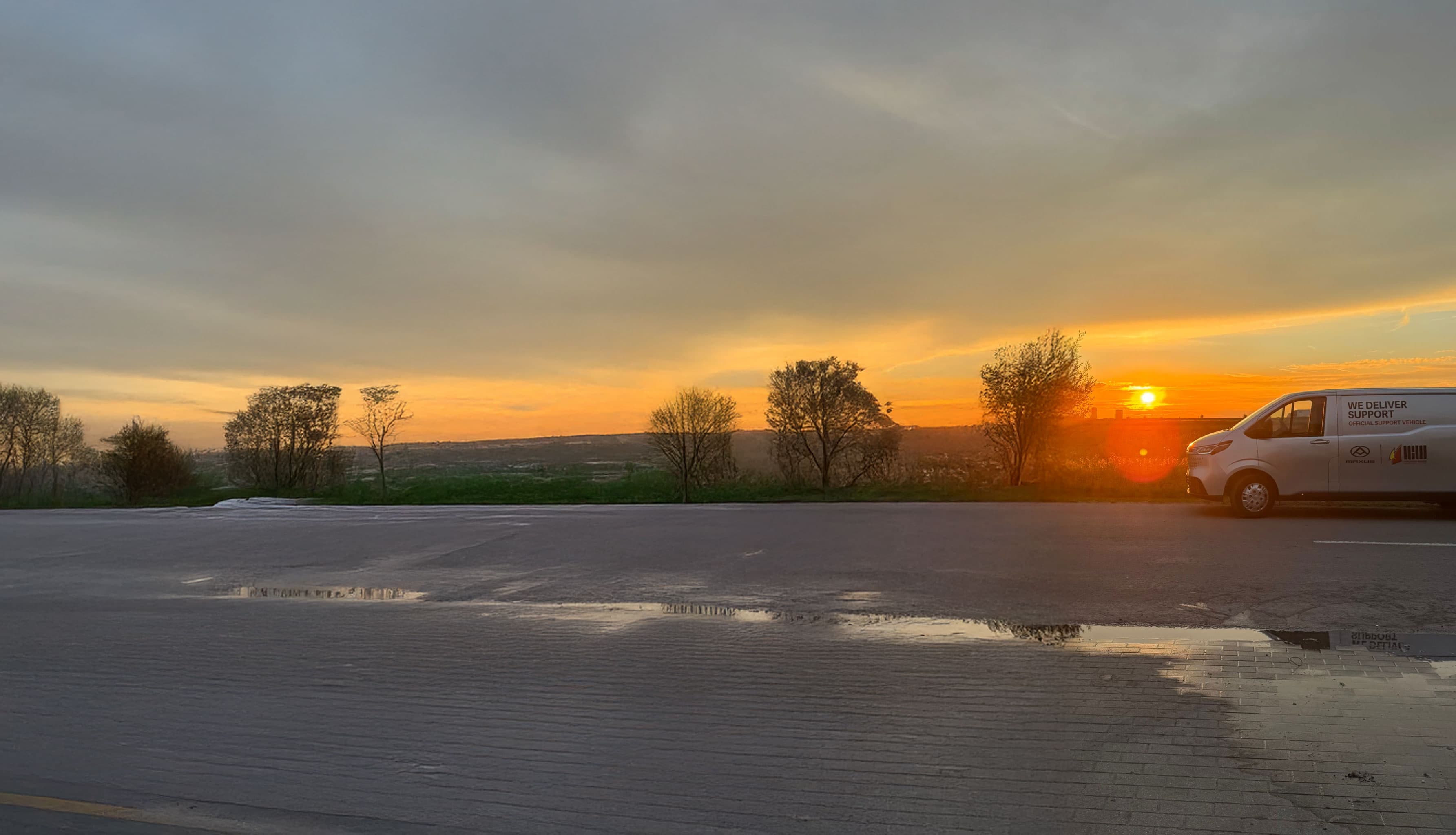 Sunset over a rural landscape with trees silhouetted against the sky, a van parked on the right, and reflections in a wet road.